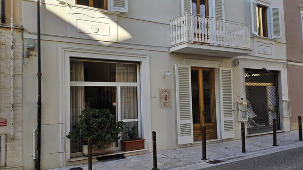 A hotel facade with a large window, a potted plant, and a door with a wooden door frame. - Verena (Montecatini Terme)