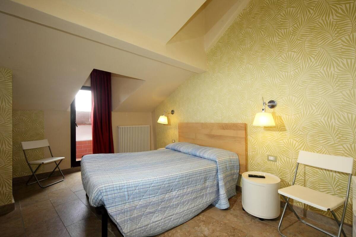 A bedroom with a bed, two bedside tables, a chair, a radiator, and patterned wallpaper.