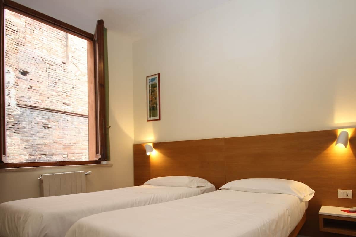 A room with two single beds, a brick wall, a radiator, and a small bedside table with a lamp.