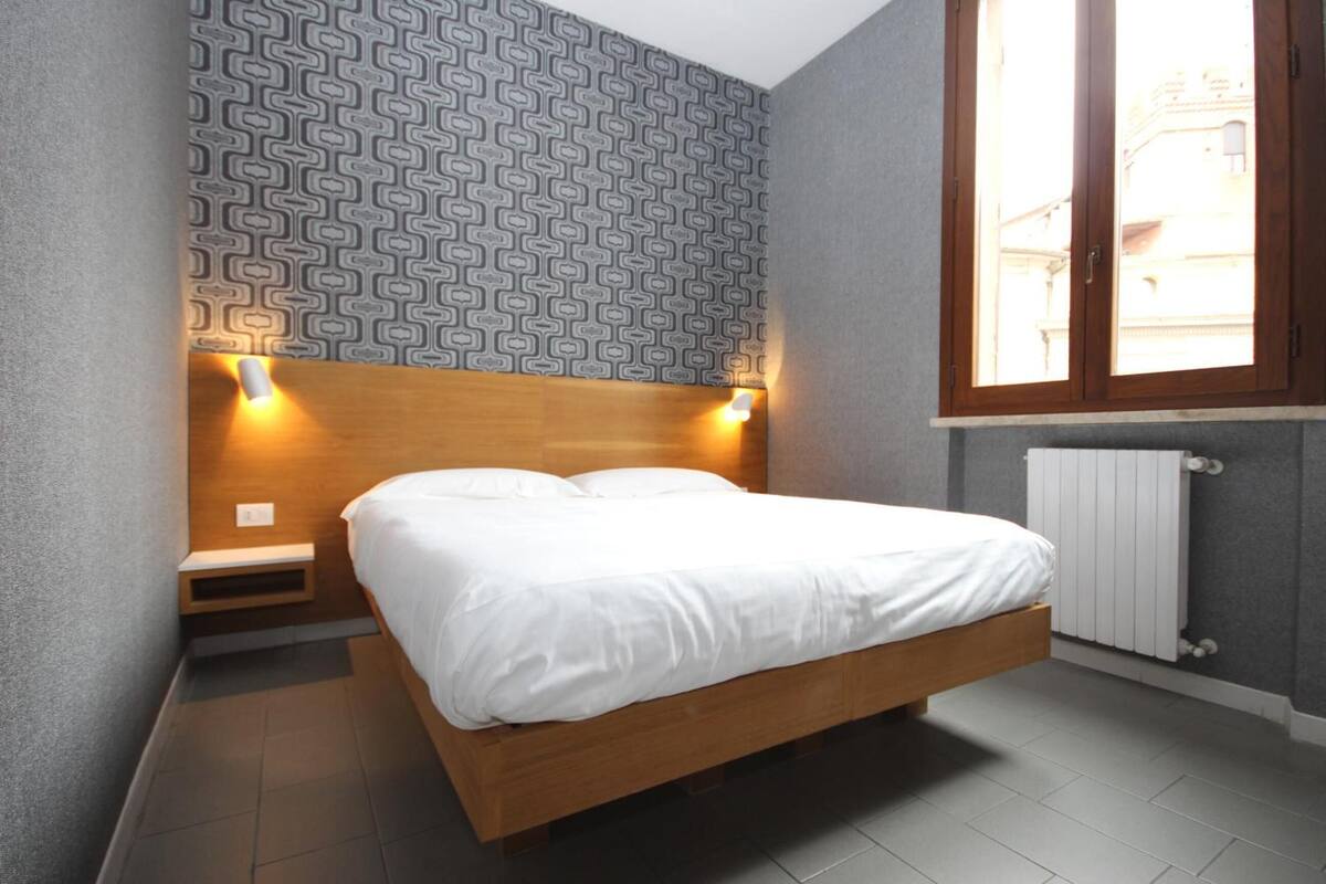 A bedroom with a wooden bed, patterned wallpaper, a window with a view, and a radiator.
