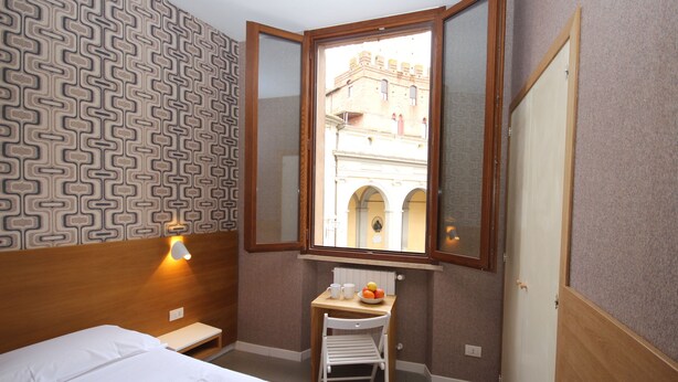 A hotel room with a bed, a small table with a bowl of fruit, a chair, a window with a view of a building, and a patterned wall. - La Perla (Siena)