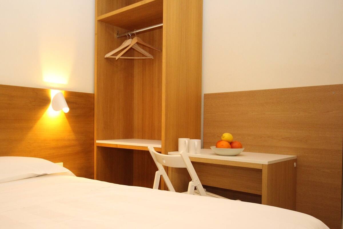 A hotel room with a bed, a desk with a bowl of fruit, a chair, and a wooden wardrobe.