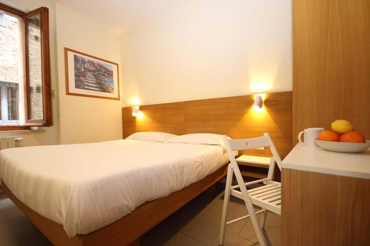 A bedroom with a wooden bed, a white chair, a small table with a bowl of fruit, and a painting on the wall.