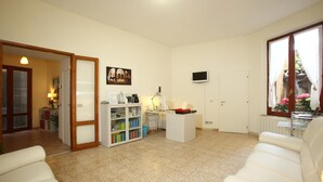Una sala de estar con un sofá blanco, una mesa de centro, un televisor montado en la pared y una puerta que da a otra habitación.