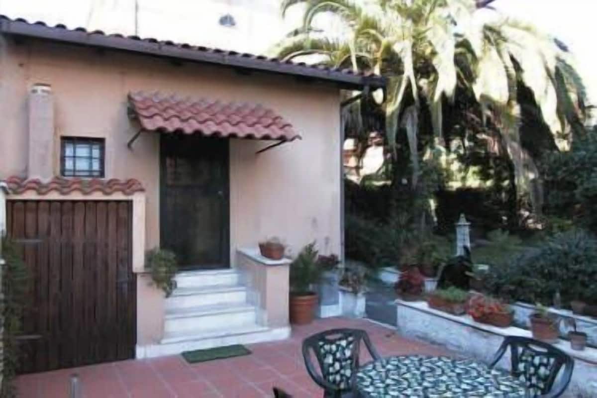 A house with a tiled roof, a wooden door, and a small porch with steps leading to the entrance. There are potted plants and a table with chairs outside.