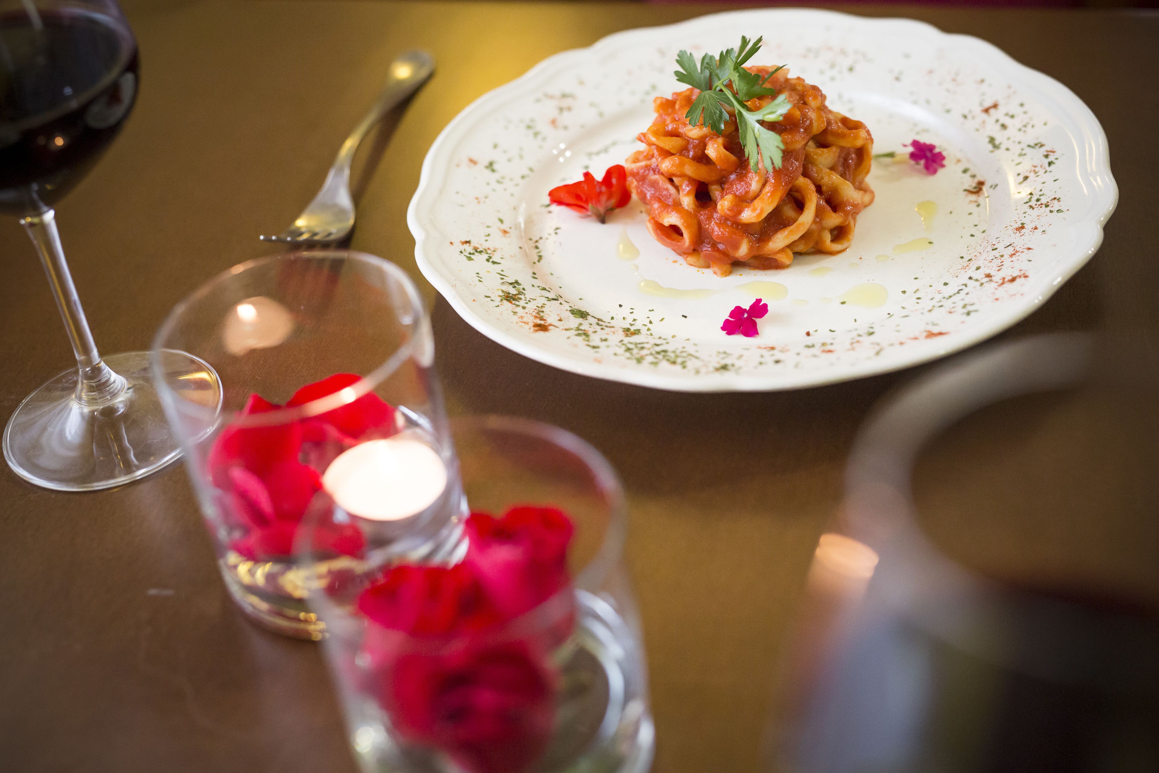 A dining table with a plate of pasta, a wine glass, and a candle with rose petals.