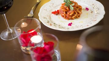 A dining table with a plate of pasta, a wine glass, and a candle with rose petals.