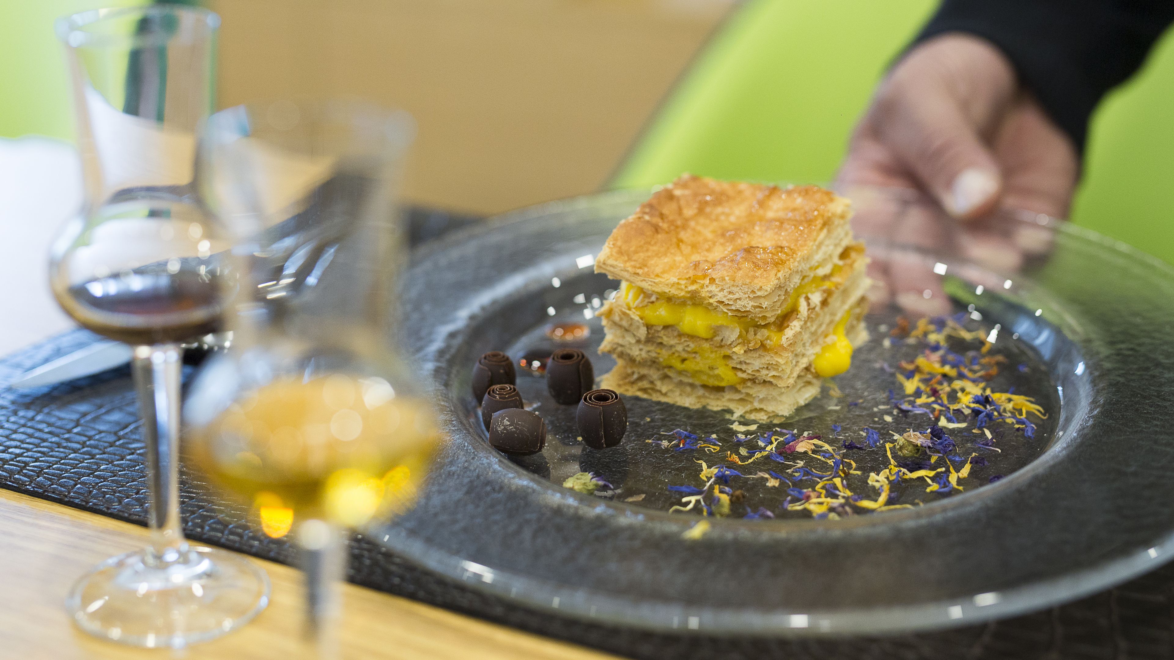 Un dessert présenté dans une assiette, agrémenté de morceaux de chocolat et de fleurs séchées, un verre de vin, et une personne floue à l’arrière-plan.