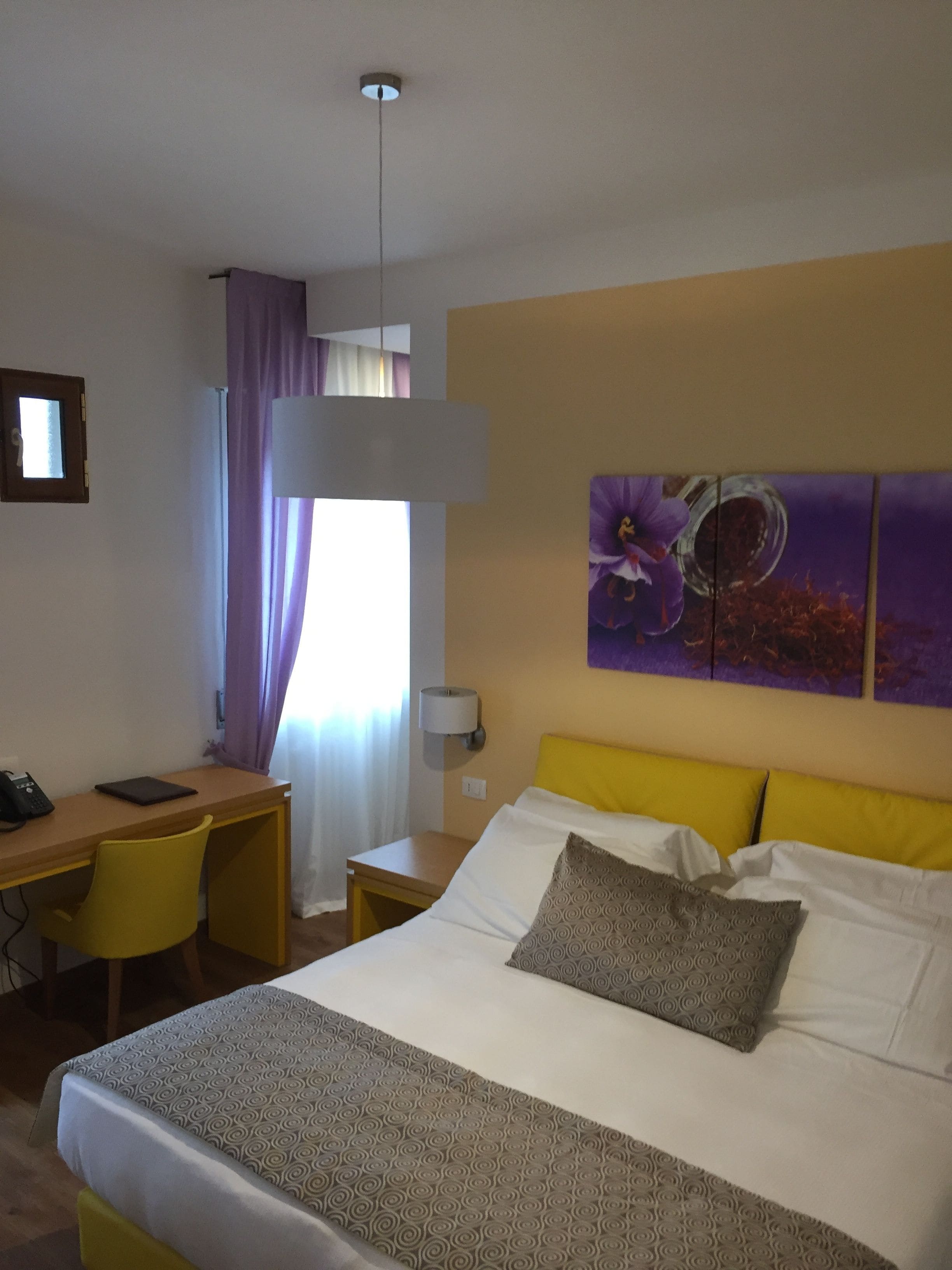 A hotel room with a bed, a desk, a chair, a lamp, and a painting on the wall.