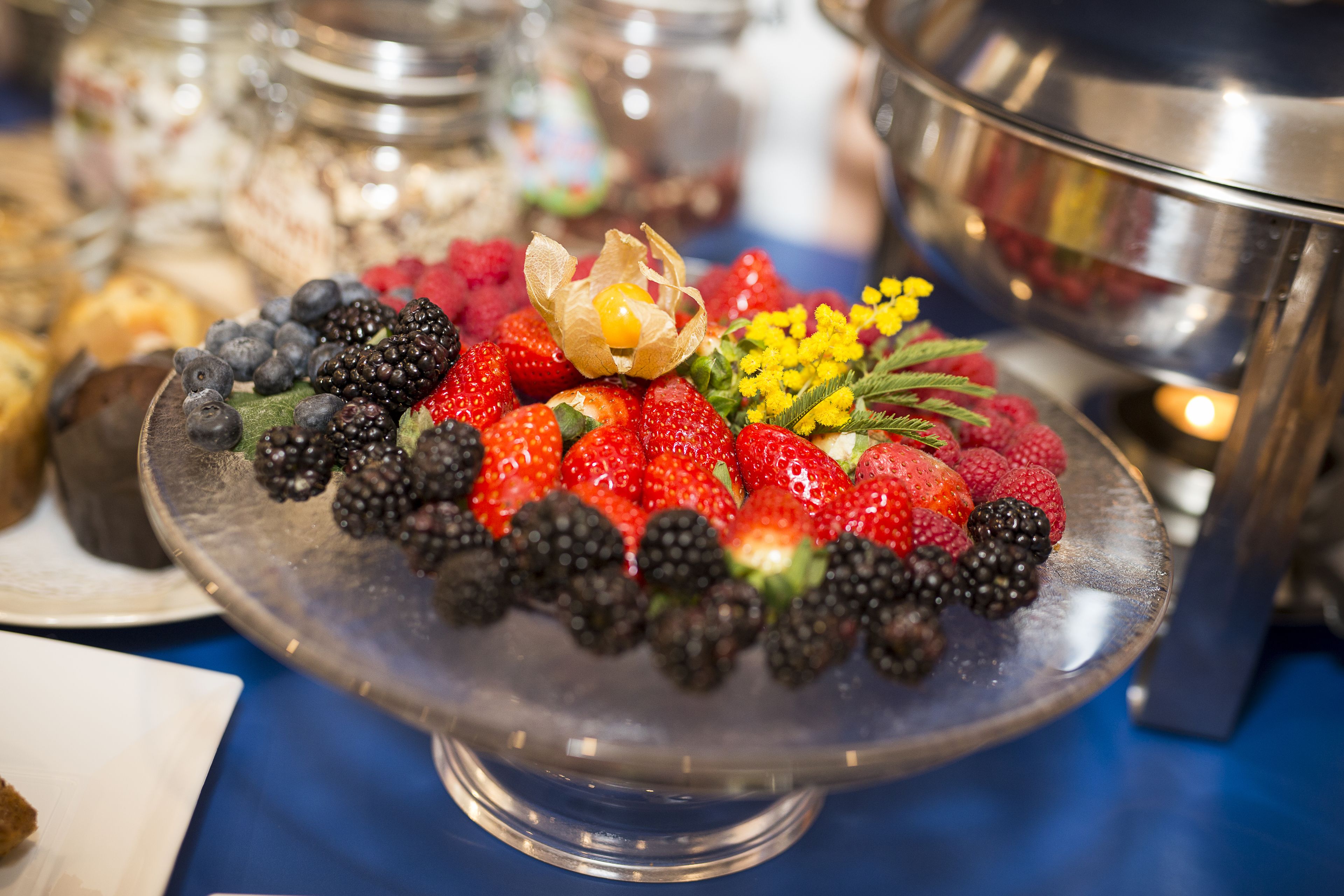 Una bandeja de frutas con fresas, moras y flores amarillas sobre un soporte de vidrio transparente.
