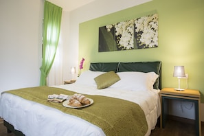 A neatly made bed with a green patterned bedspread, a plate of croissants, and a green pillow. There is a wall-mounted picture of white flowers, a bedside lamp, and a small table with a lamp. - La Gustea Hotel & Cucina (Sarteano)