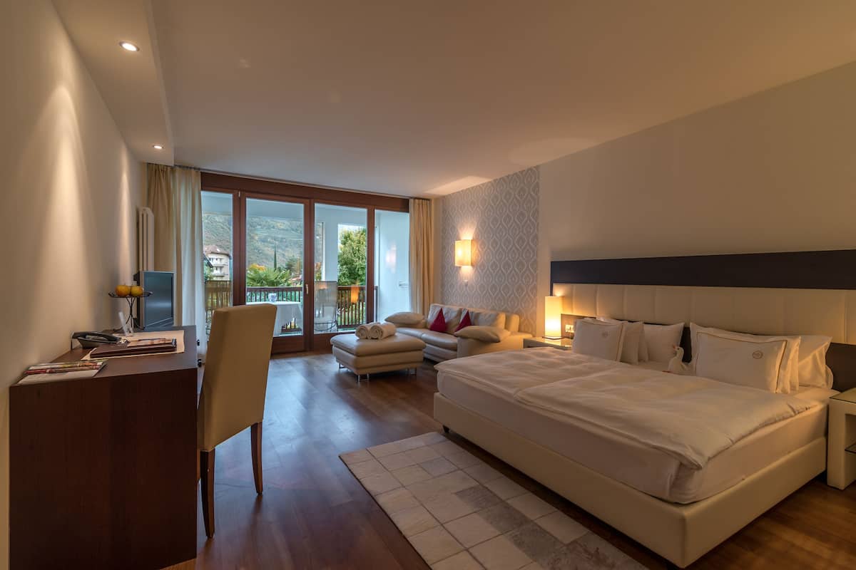 A modern hotel room with a large bed, a desk, a chair, a sofa, and a balcony with a view of the mountains.