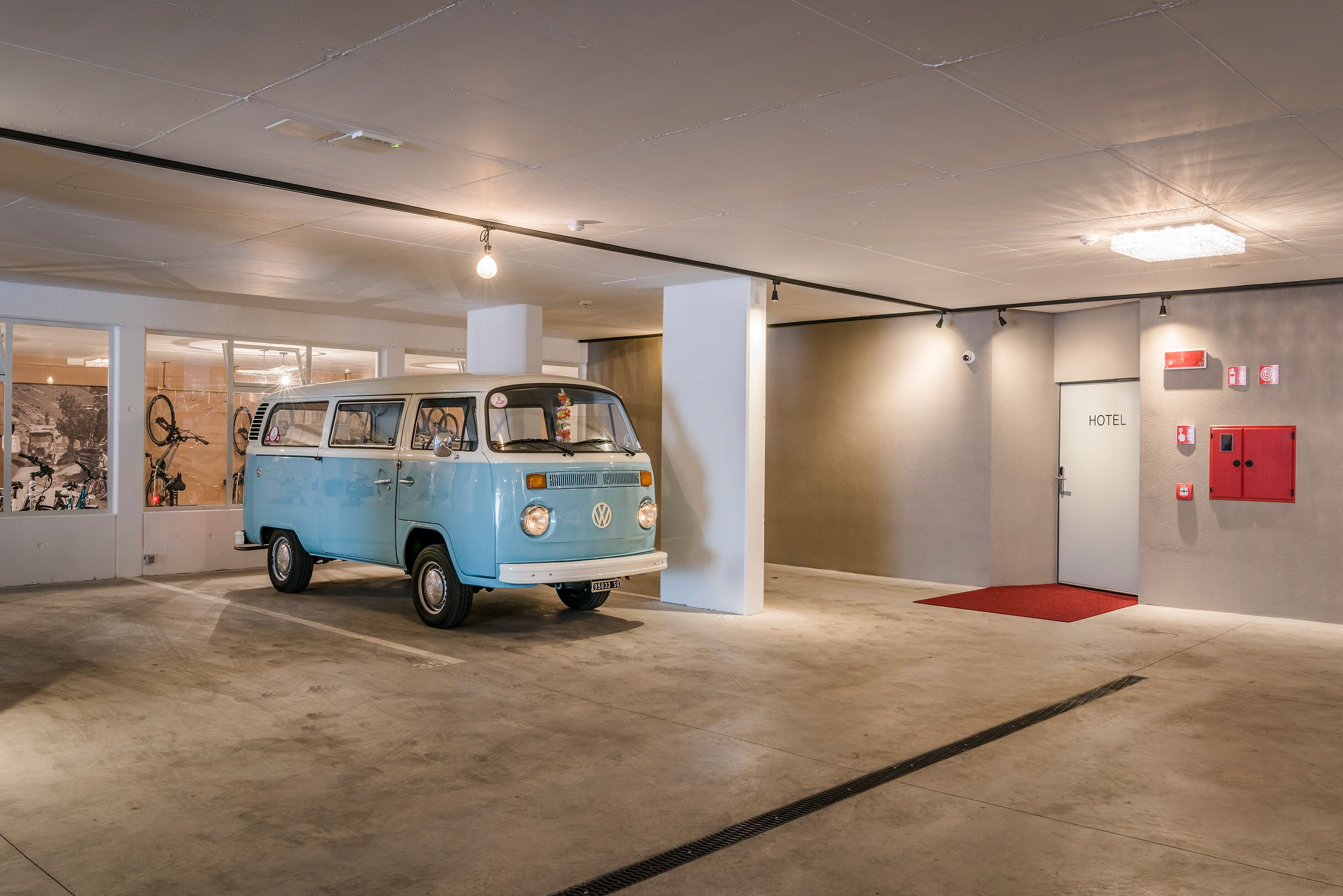 A vintage blue van parked in a modern hotel lobby with a red carpet, a visible door labeled "HOTEL", and a wall-mounted fire alarm.
