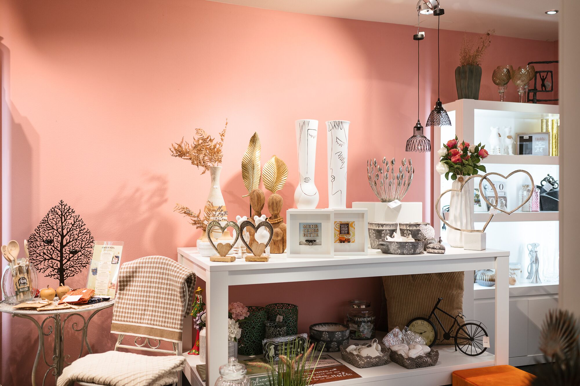 A well-organized room with a pink wall, a white console table displaying various decorative items, a chair, and a bookshelf filled with books and decorative objects.