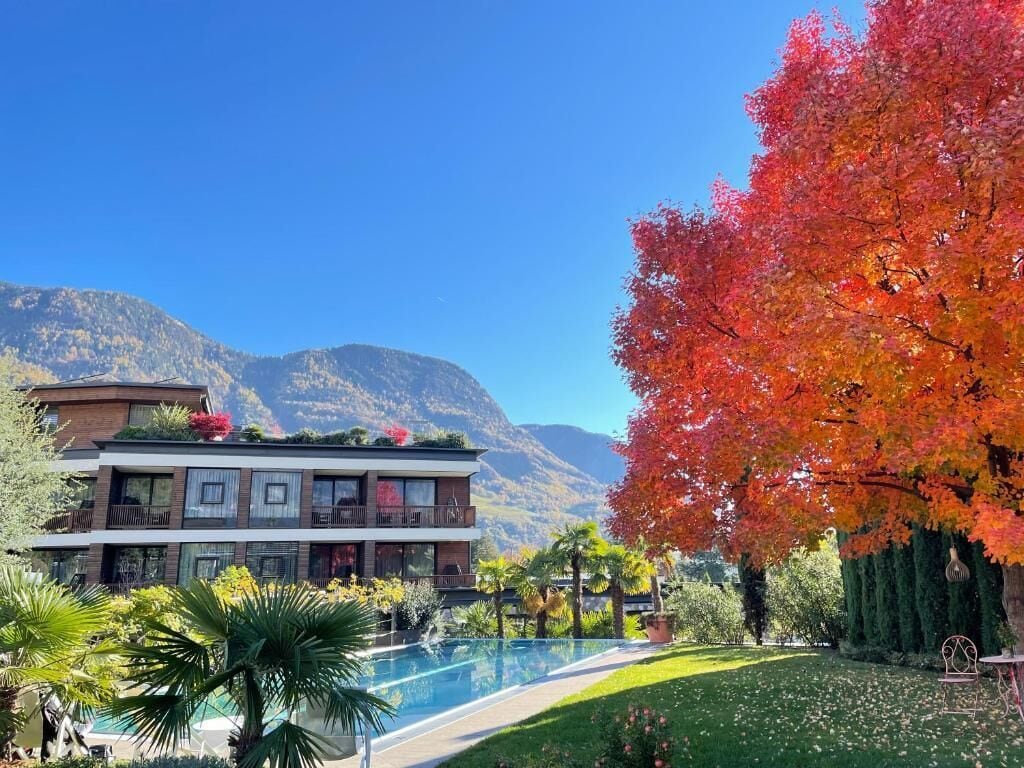 Ein modernes Hotel mit Pool, umgeben von üppiger Vegetation und herbstlich gefärbten Bäumen, vor einer Bergkulisse gelegen.