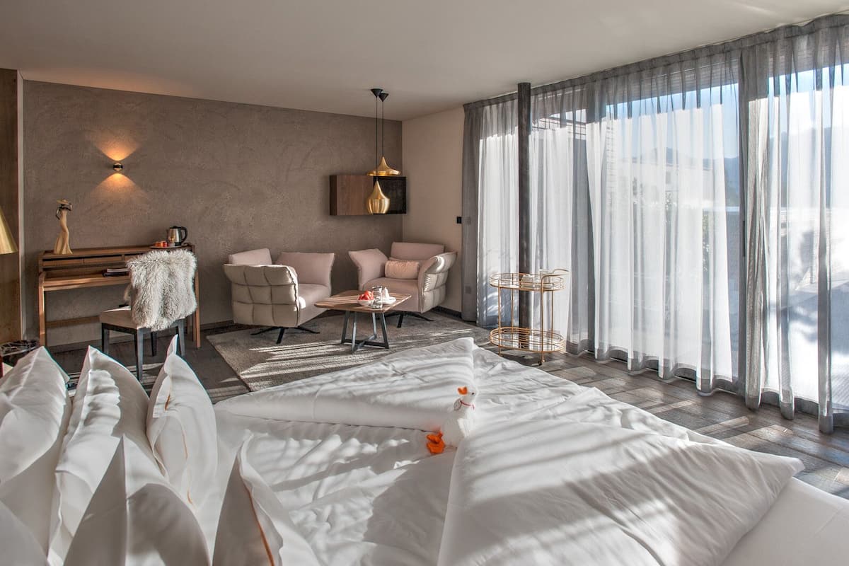 A modern hotel room with a large bed, a sitting area with chairs, a small table, and a piano.
