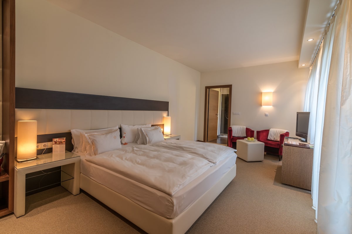 A hotel room with a large bed, bedside tables, a desk, a red armchair, a TV, and a mirror.