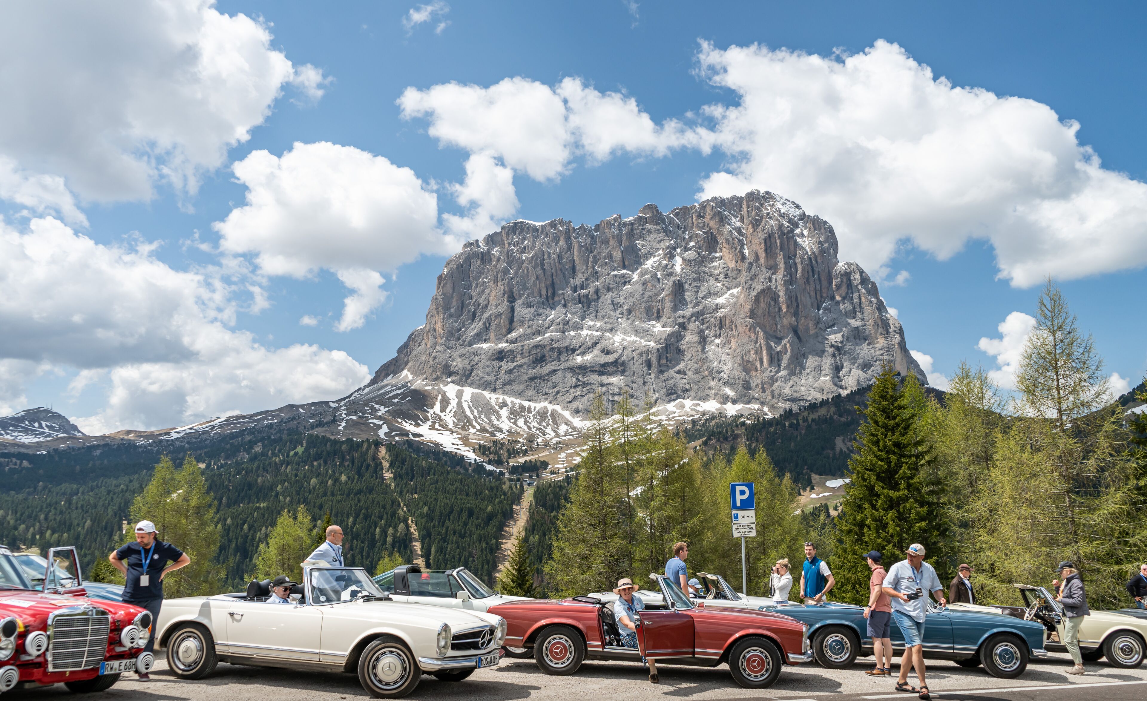 Een parkeerplaats met oldtimers en een bergachtig landschap op de achtergrond.