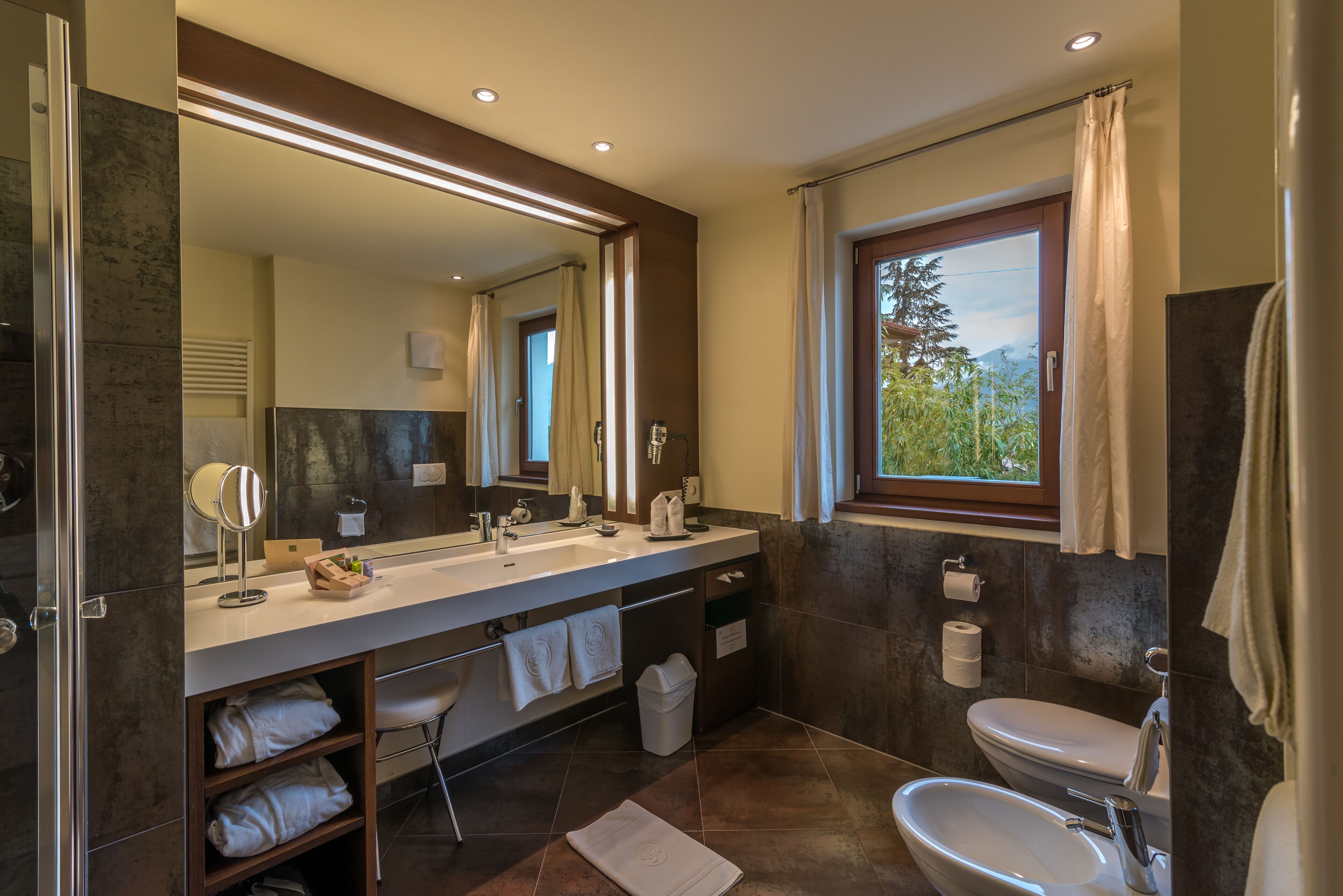 A modern bathroom with a large mirror, dual sinks, and a window with a view of greenery.