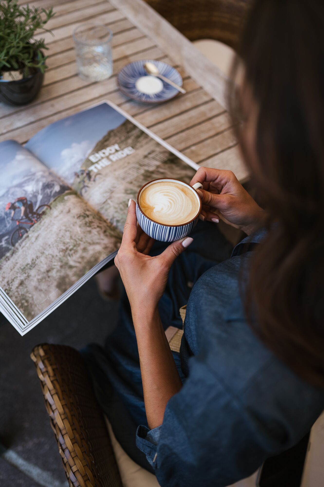 Una persona sosteniendo una taza de café, sentada a una mesa con una revista y una planta.
