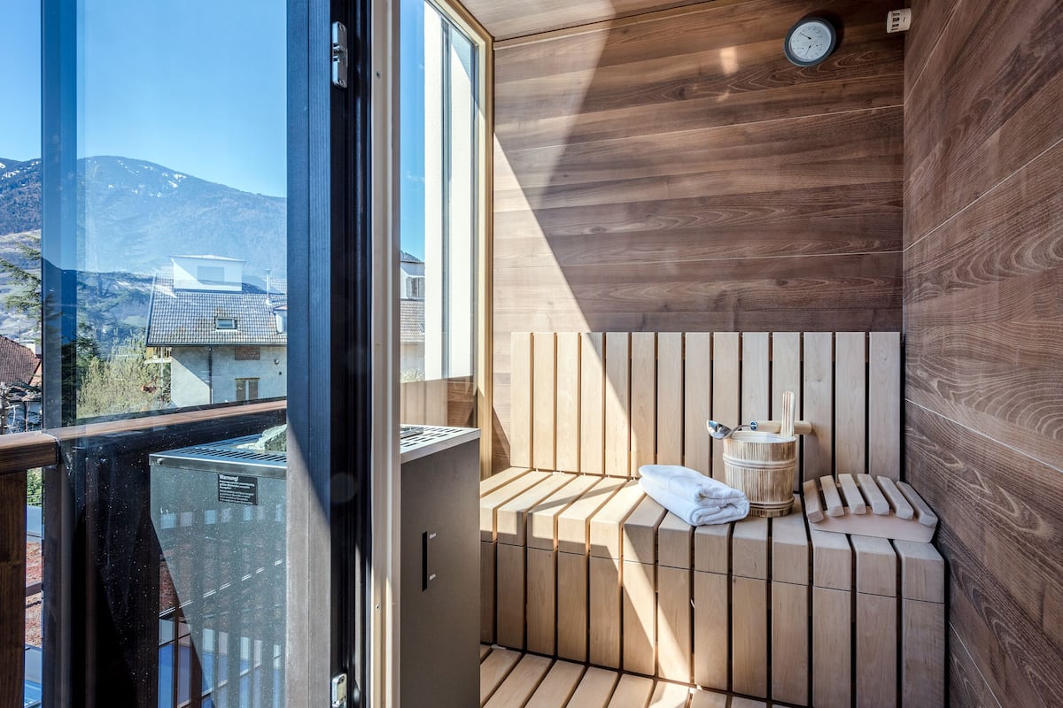 A modern sauna with wooden slats, a towel, and a wooden bucket, with a view of mountains through a large window.