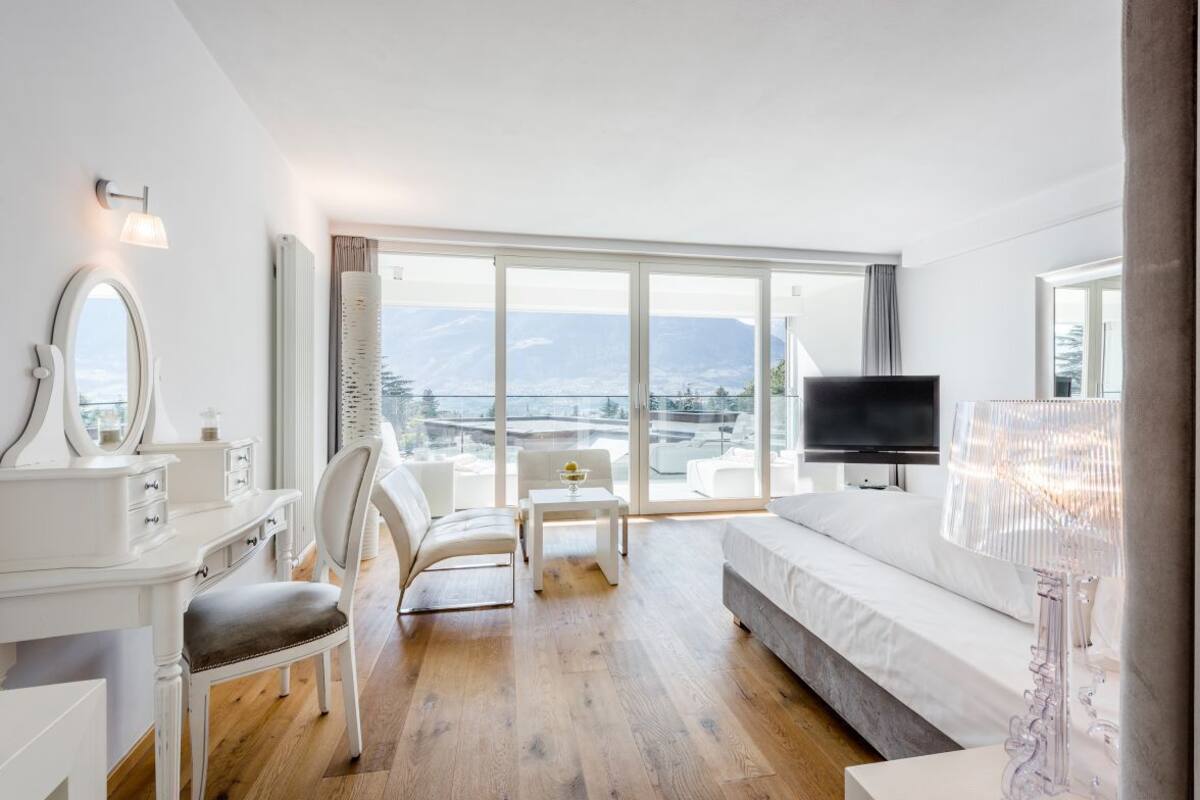 A modern hotel room with a large window, a bed, a TV, a desk with a mirror, and a chair.