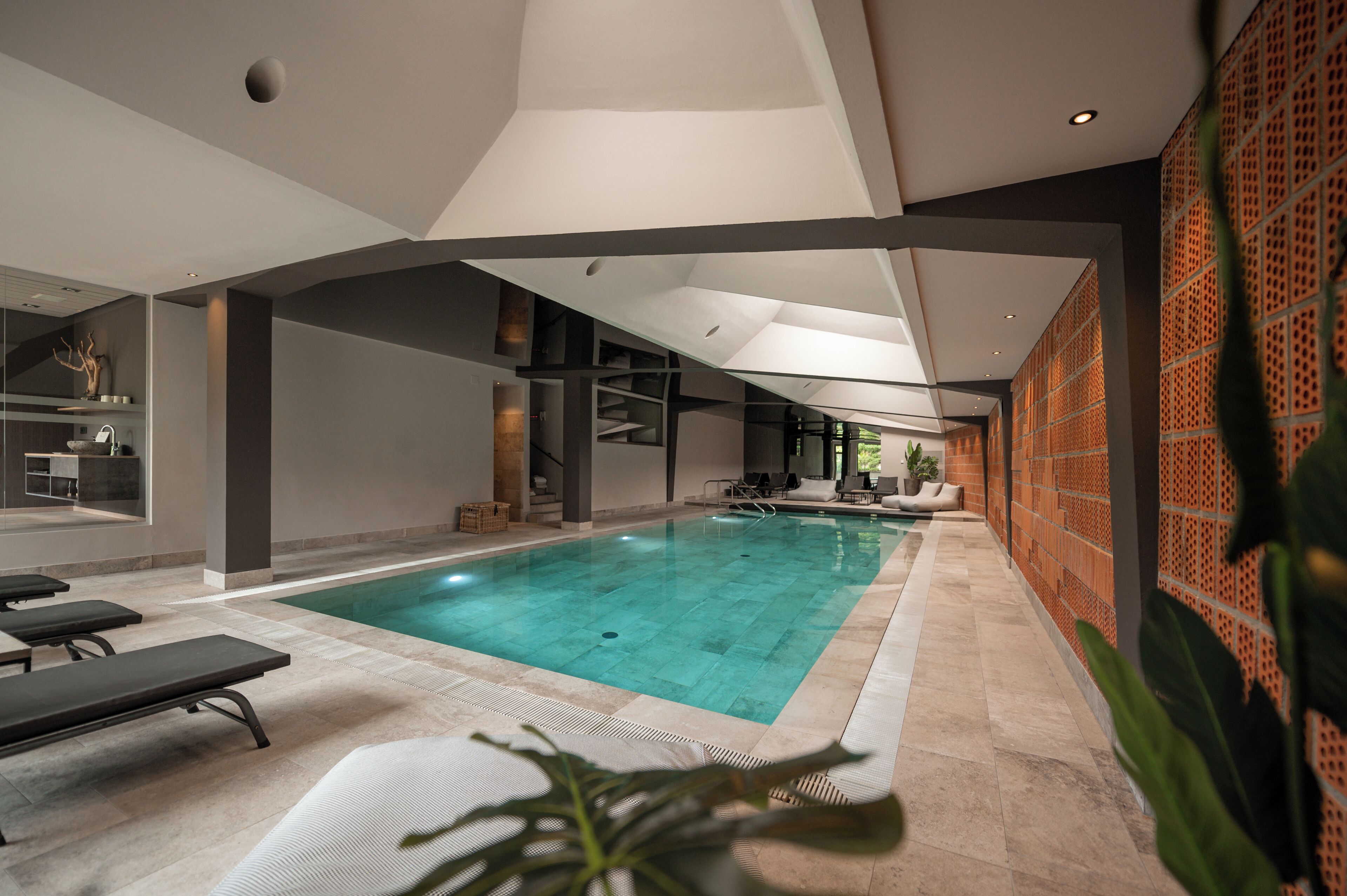 A modern indoor swimming pool with clear water, surrounded by lounge chairs and a brick wall with a lattice design.