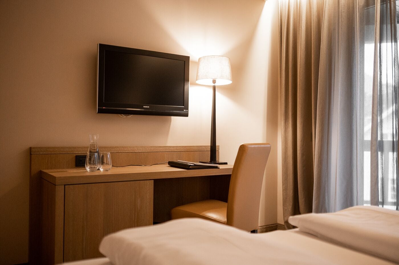 A hotel room with a bed, a desk with a lamp, a TV, and a chair.