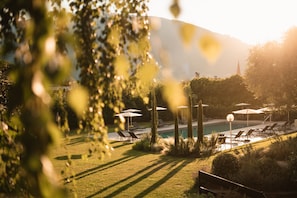 A pool area with lounge chairs and umbrellas, surrounded by greenery and a mountain in the background. - Pollinger (Merano)