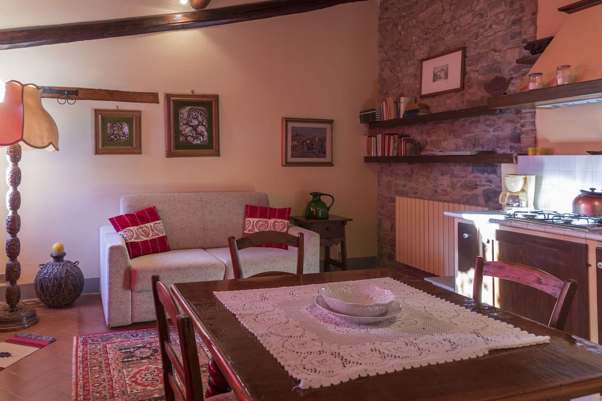 Ein gemütliches Wohnzimmer mit einer Couch, einem Esstisch mit weißer Tischdecke und einer Küchenzeile mit Herd und Regalen.