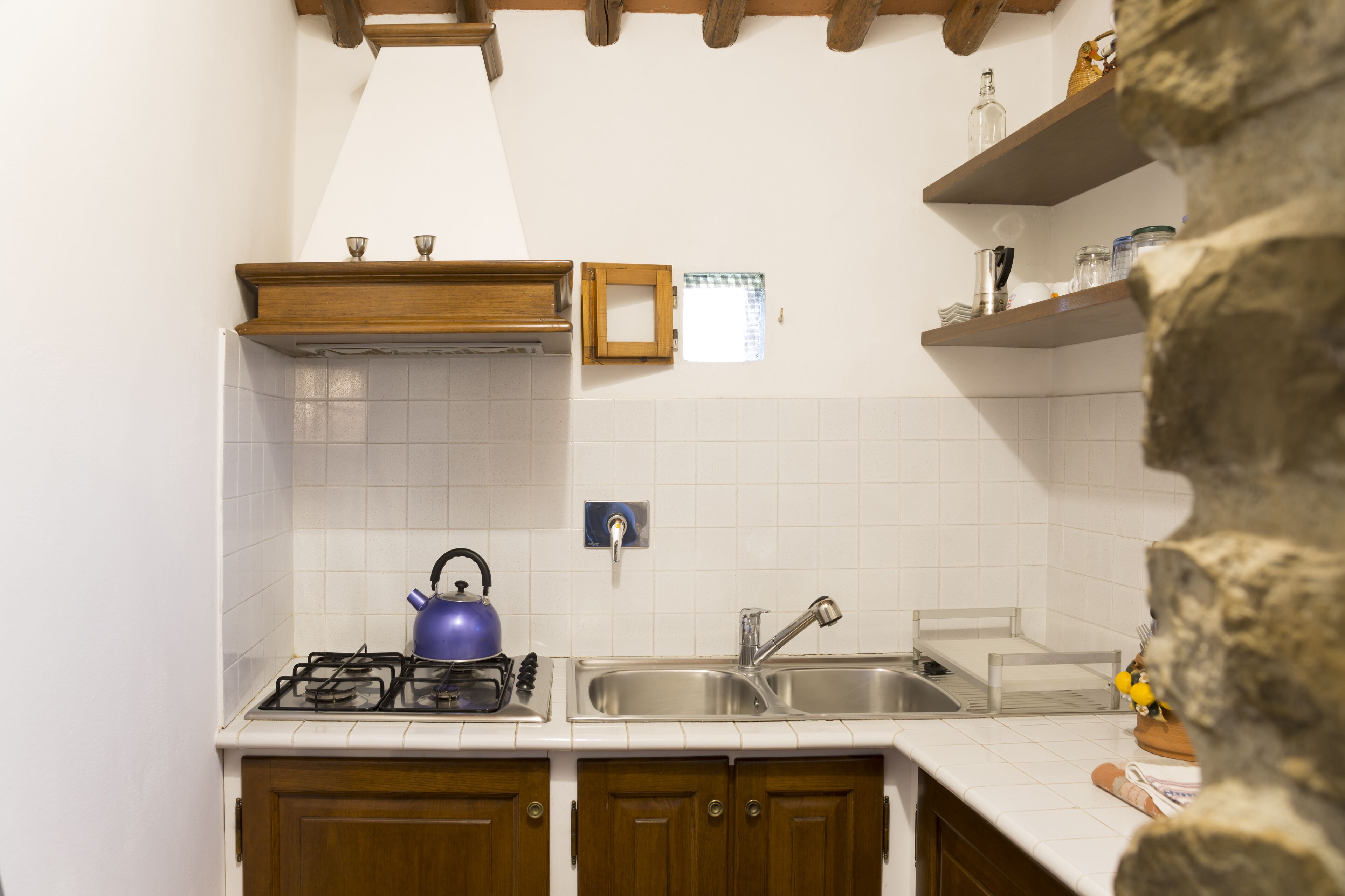 A kitchen with white tiled walls, wooden cabinets, a gas stove, a sink, and a stone wall.