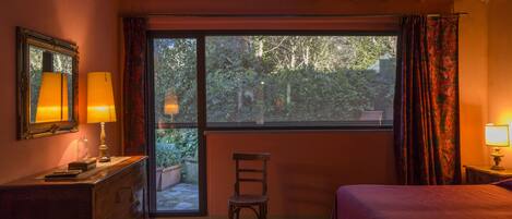 Un dormitorio con cama, una silla, un aparador, una ventana con cortinas y una lámpara.