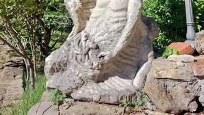 Een stenen beeld van een figuur met een halo, omringd door groen en een lantaarnpaal.
