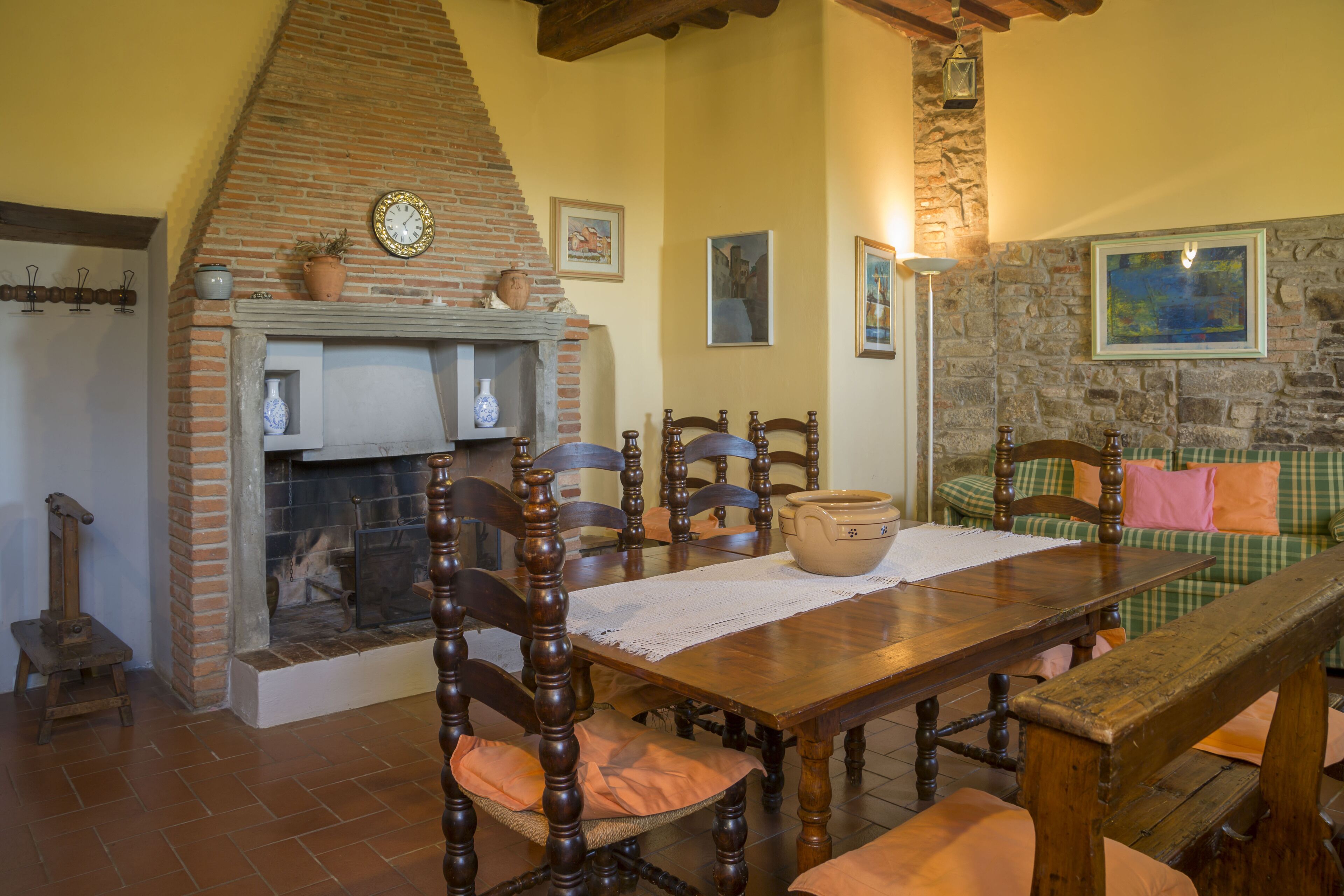 A dining area with a brick fireplace, wooden chairs, a wooden table, and a stone wall with a painting.