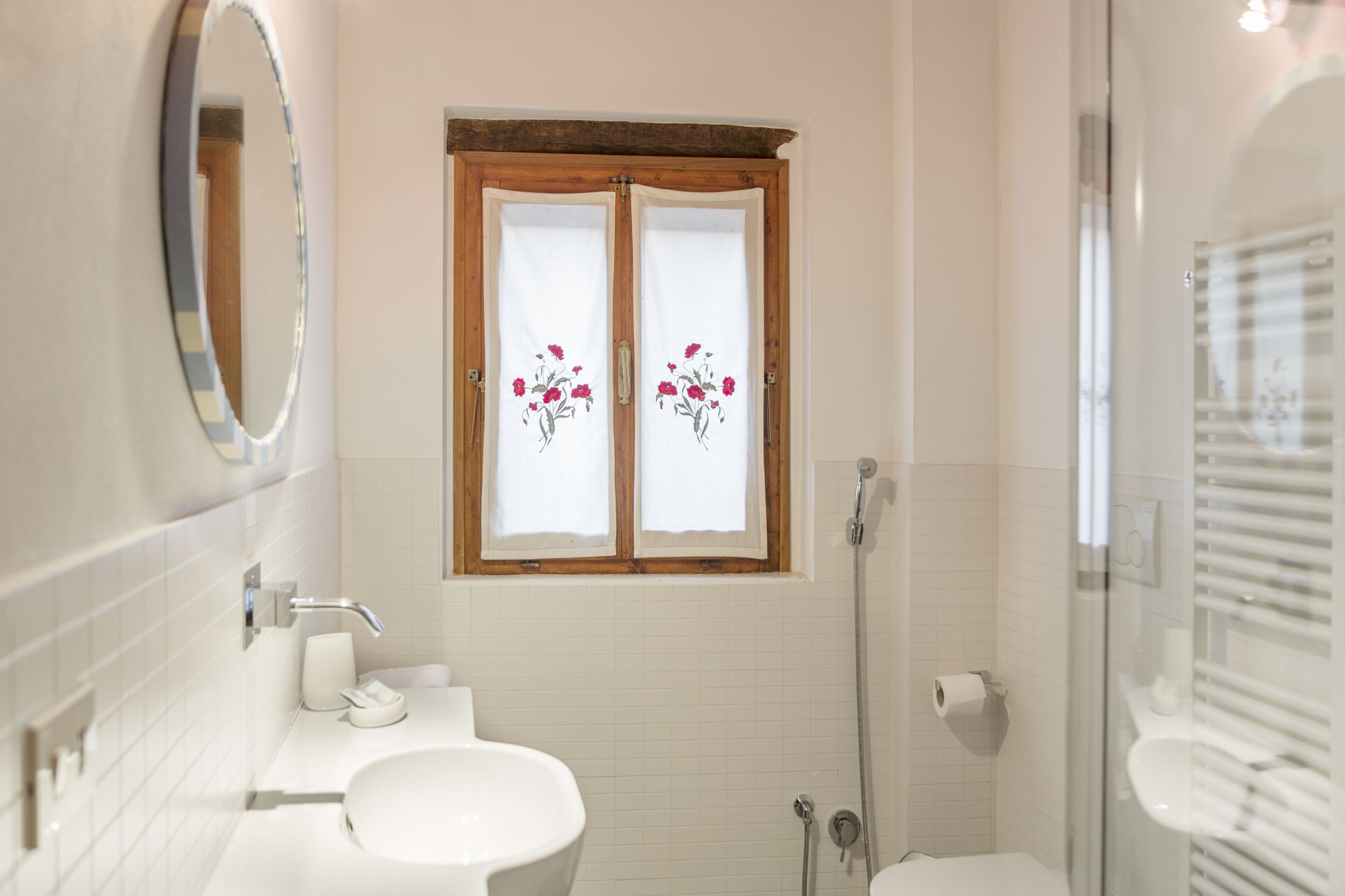 A bathroom with a white sink, a mirror, a window with floral curtains, and a shower.