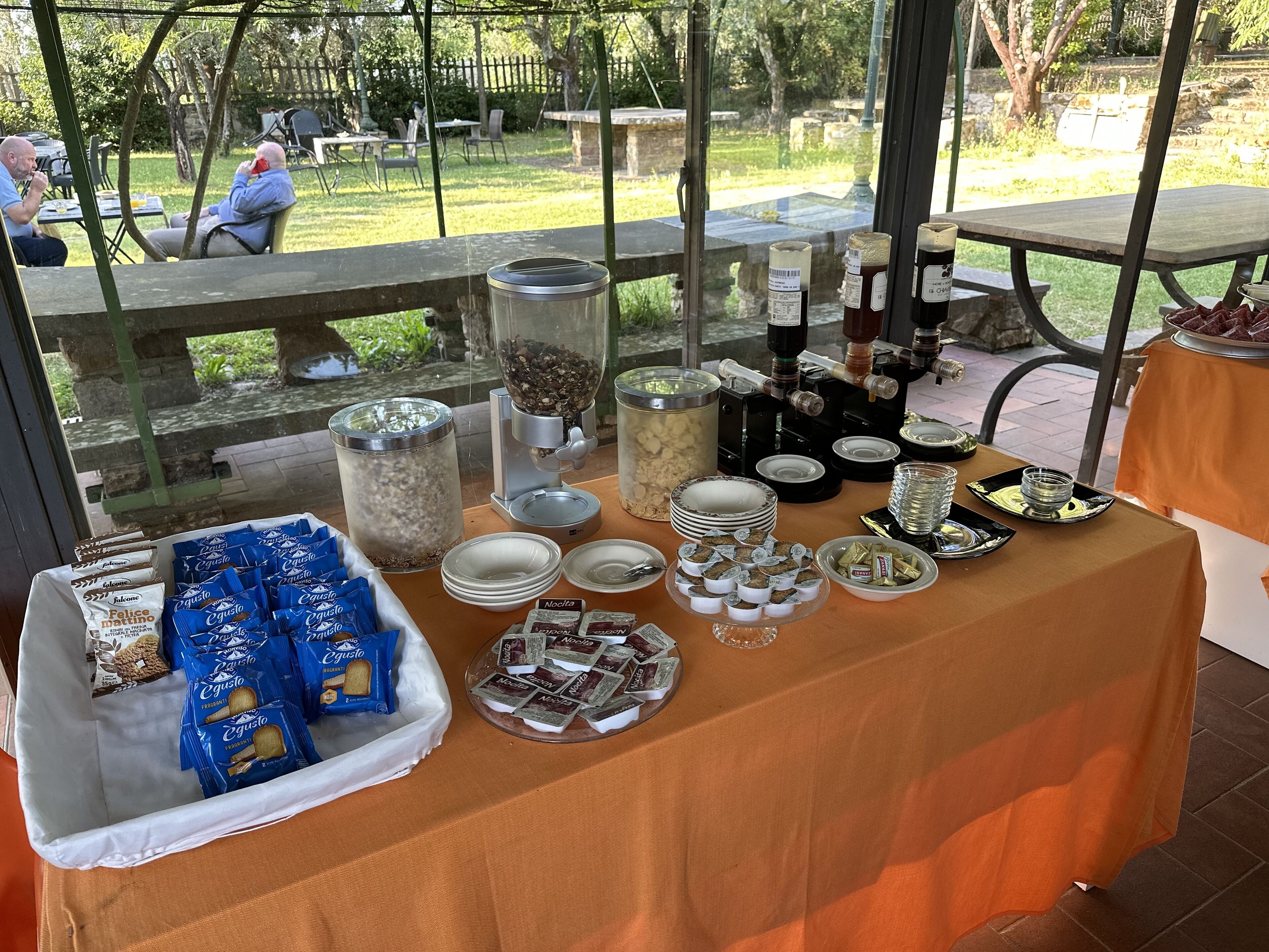 Une table de buffet proposant divers plats et accompagnements, installée dans un espace extérieur avec vue sur un jardin et une terrasse.