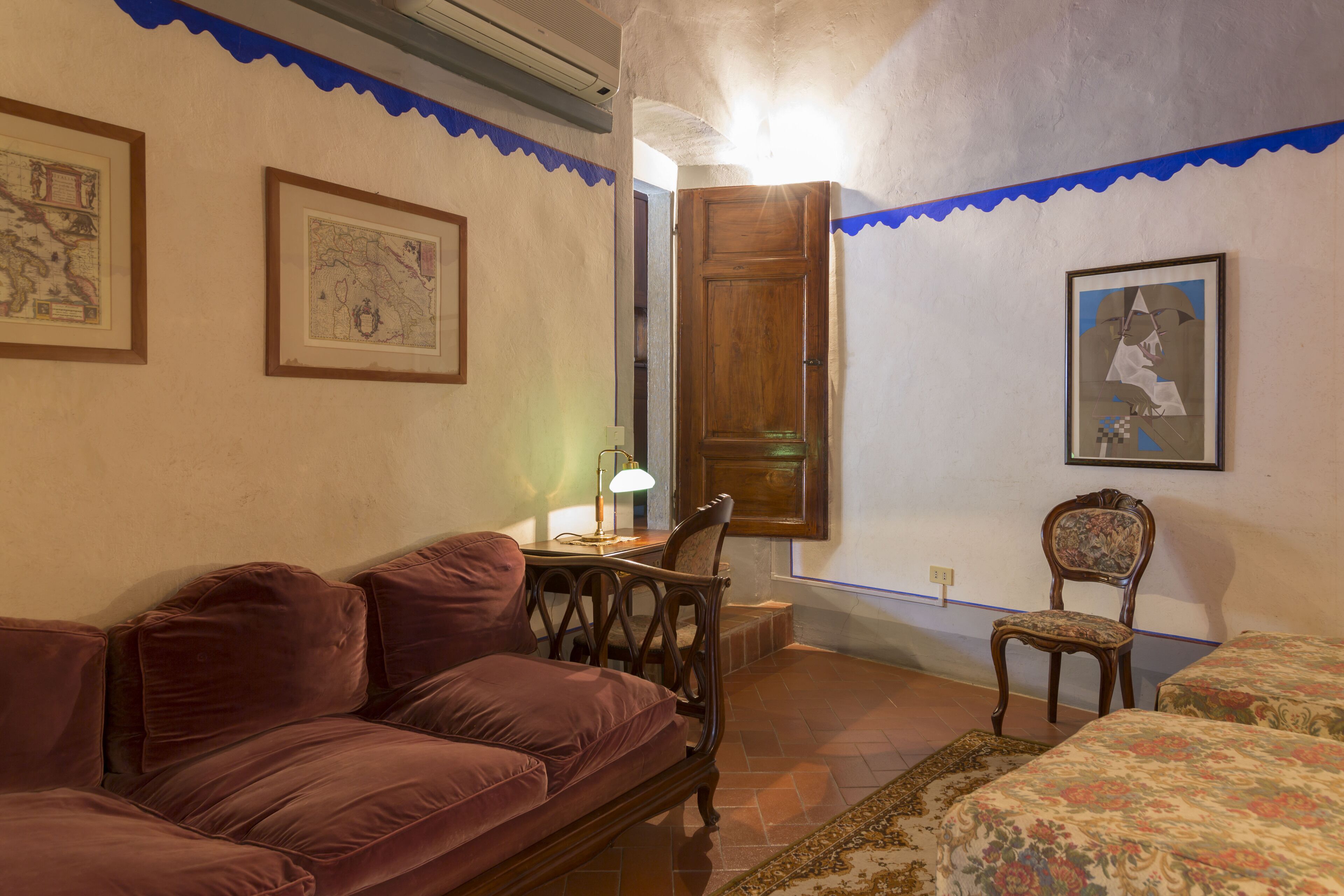 Quarto com sofá de couro marrom, uma cadeira de madeira, uma mesinha com abajur e cama com roupa de cama floral.