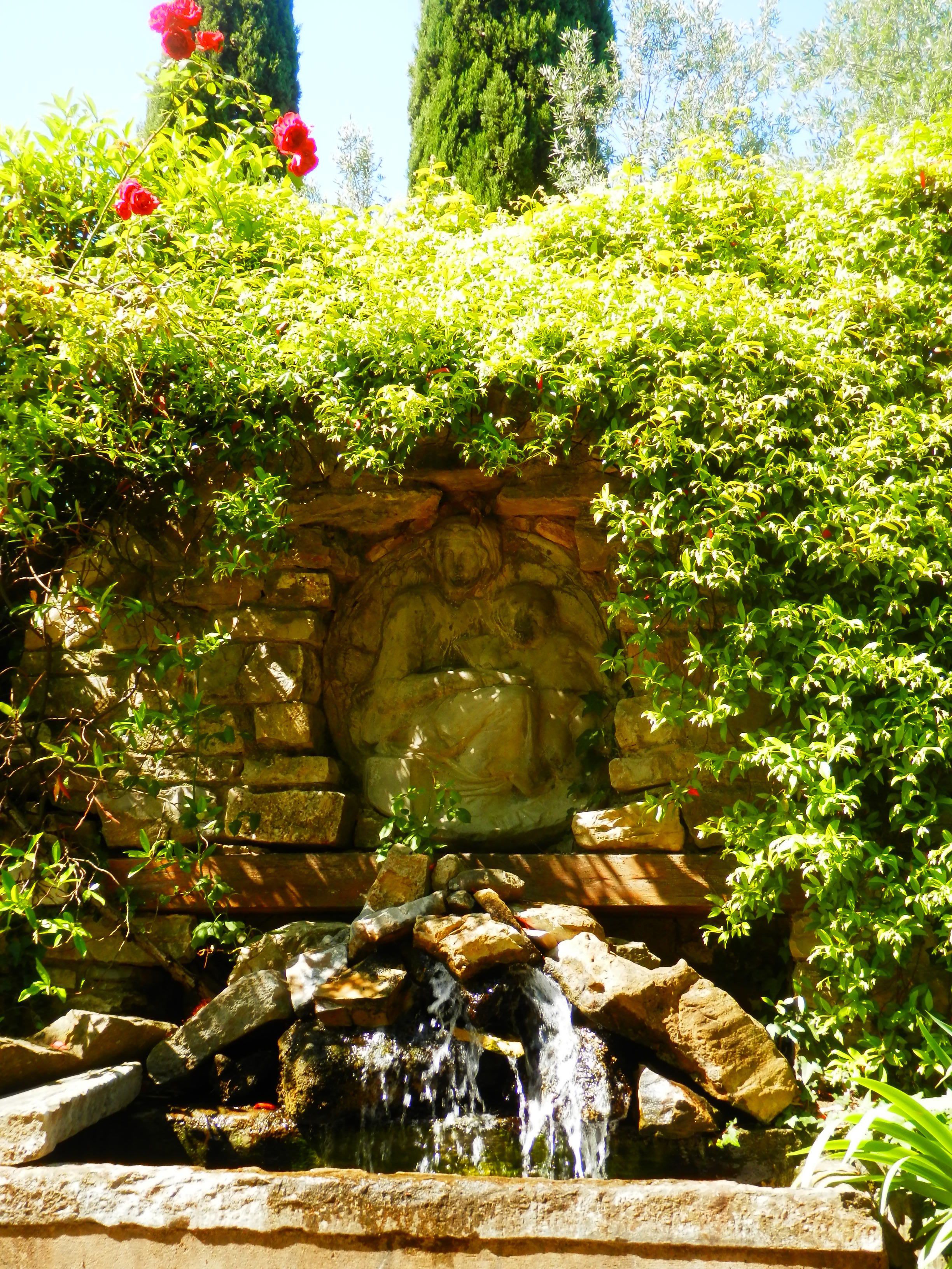 Een stenen muur met een gebeeldhouwde figuur, een kleine waterval en weelderige begroeiing.