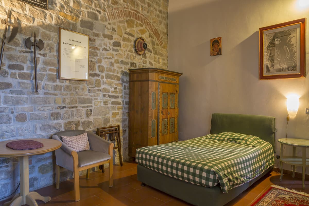Ein Zimmer mit Steinwand, einem Bett mit kariertem Überwurf, einem Holzschrank, einem Stuhl und einem kleinen Tisch.