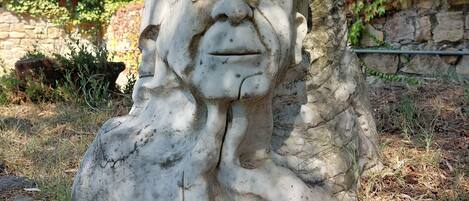 Una escultura de piedra de una cabeza humana con barba, ubicada en un jardín con un muro de piedra y árboles al fondo.