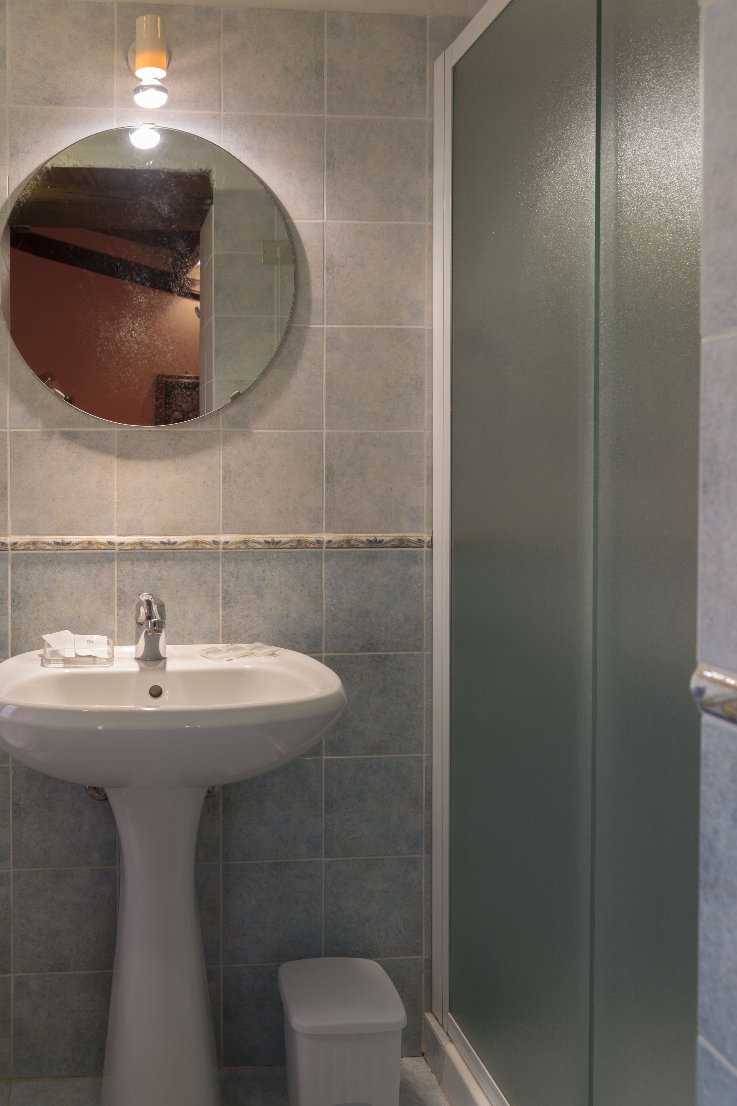A bathroom with a pedestal sink, a round mirror, and a glass shower enclosure.