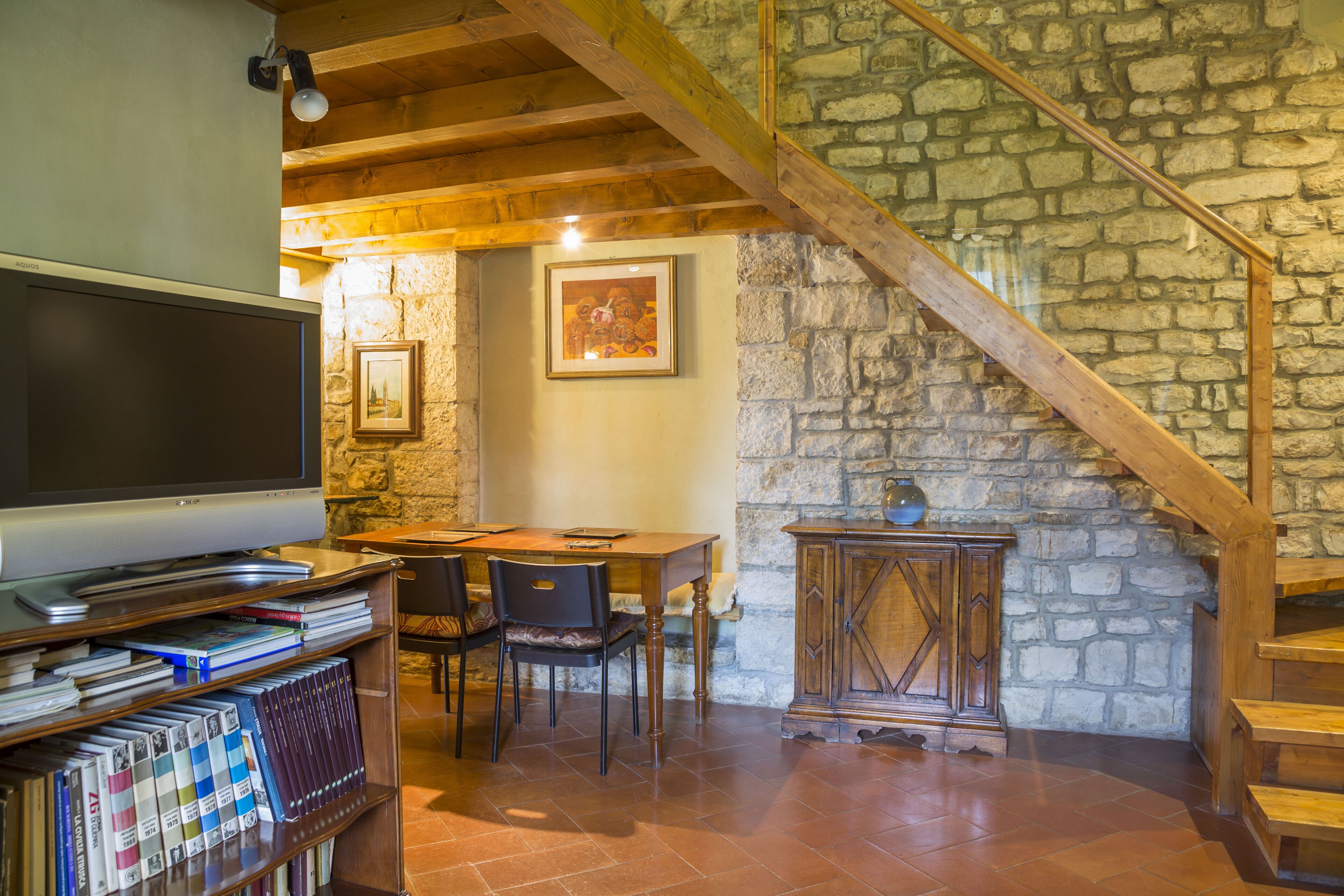 A cozy living room with a television, bookshelves, a dining table, chairs, and a stone wall.