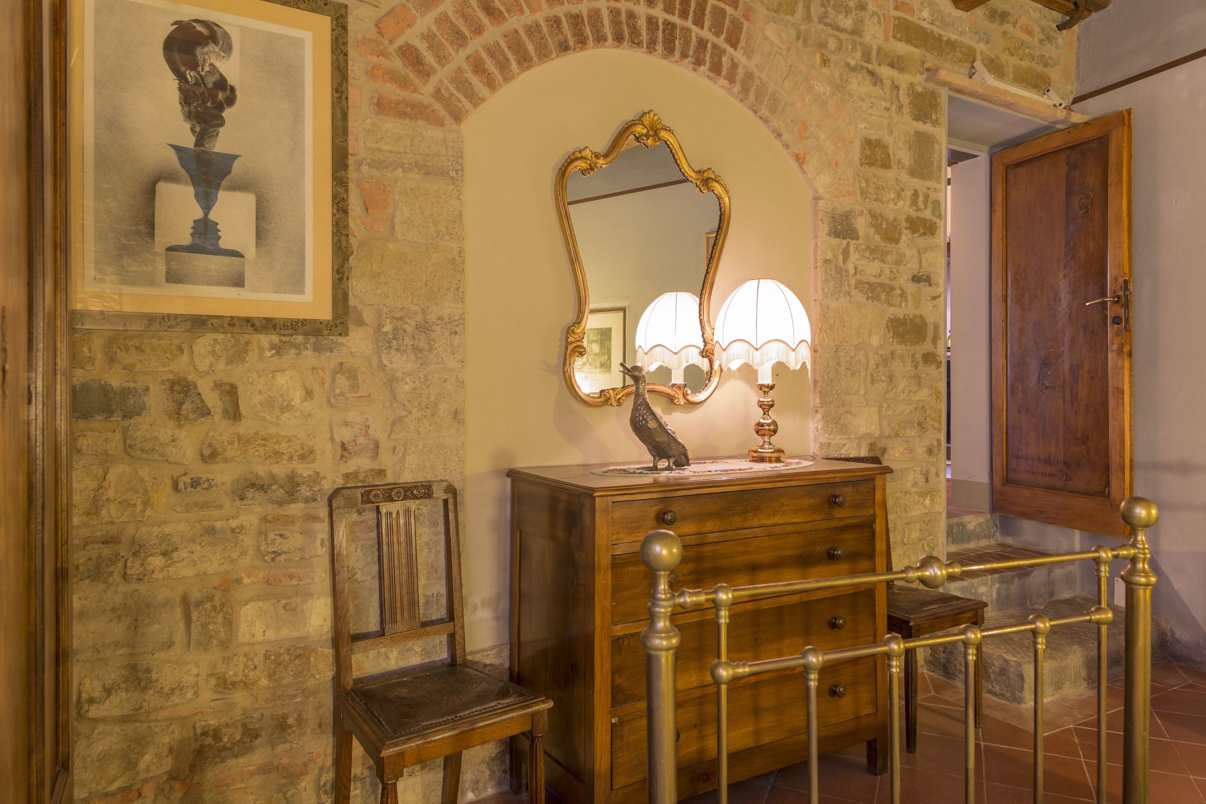 Una habitación con pared de piedra, un aparador de madera con cajones, una silla, un espejo con marco decorativo y una puerta de madera.