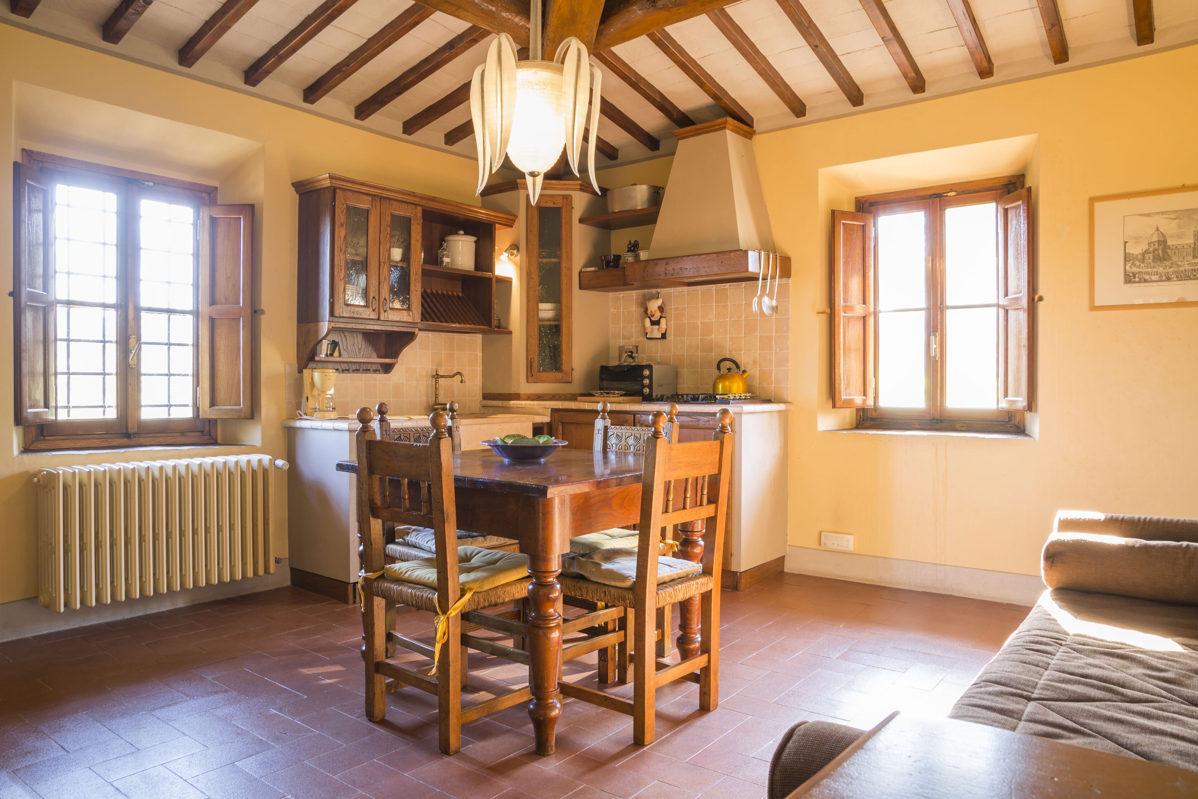 A spacious kitchen with wooden cabinets, a dining table, and chairs. There is a window providing natural light and a radiator under the window.