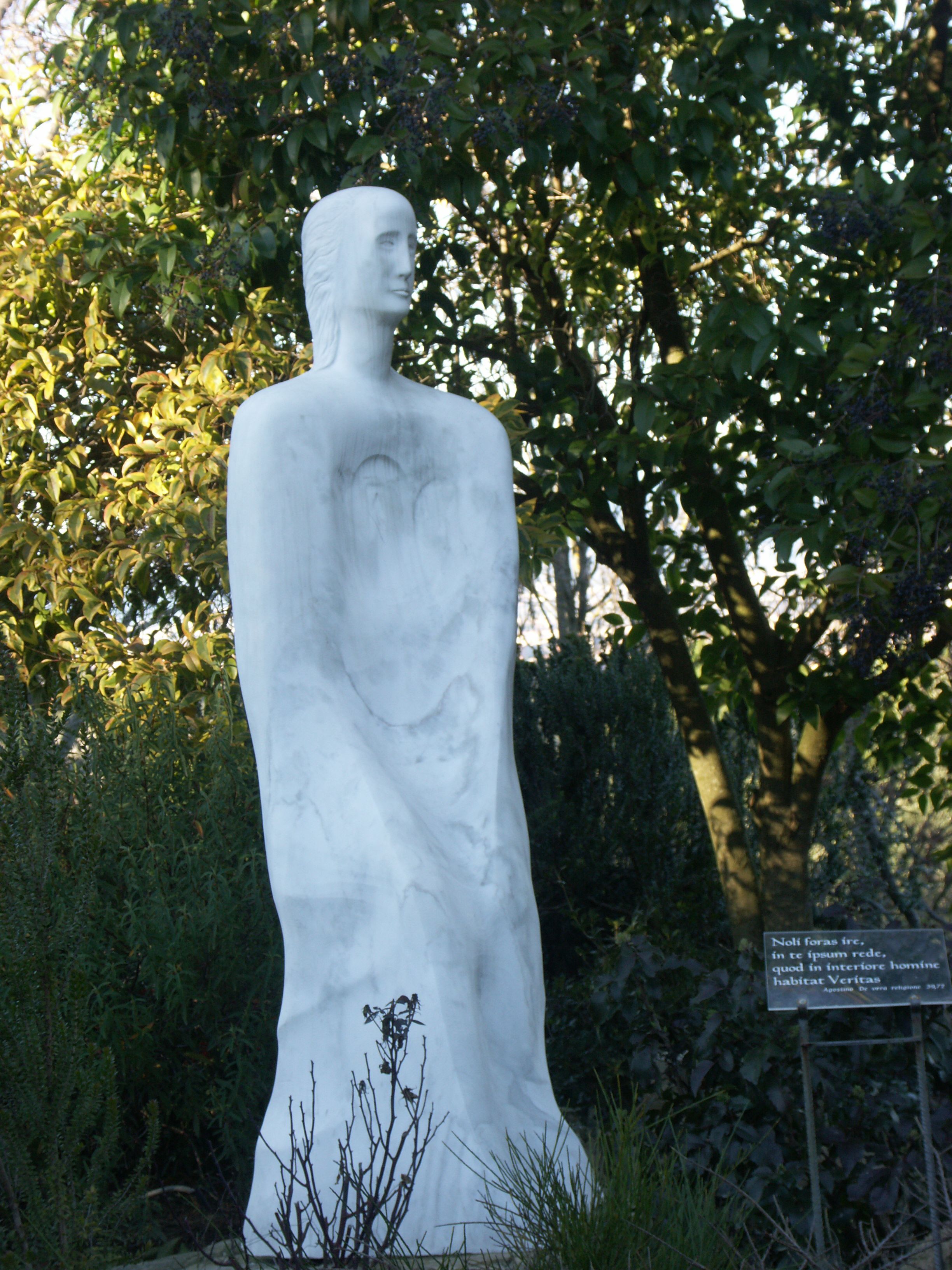 A white statue of a woman with a child in her arms, set against a backdrop of trees and foliage.