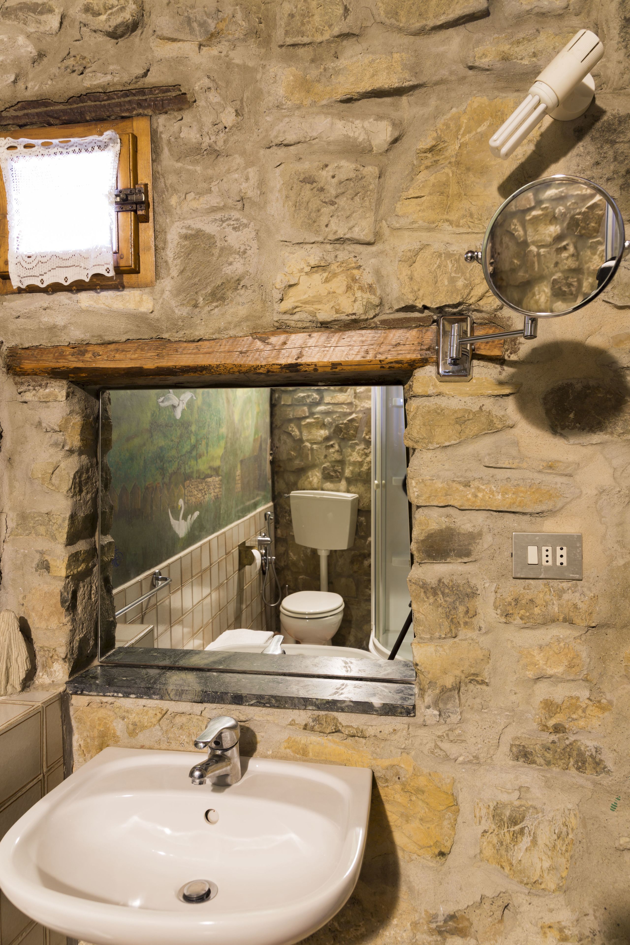Une salle de bain avec un mur en pierre, un lavabo blanc, un miroir et une fenêtre donnant sur une forêt.