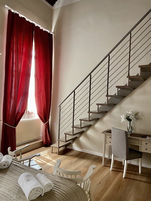 A bedroom with a wooden floor, a bed with white linens, a desk with a chair, a staircase with metal railings, and a window with red curtains. - Soggiorno Alessandra (Florence)