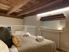 A bedroom with a wooden ceiling, a white metal bed frame, a beige bedspread, and a mirror with a light above it. - Soggiorno Alessandra (Florence)