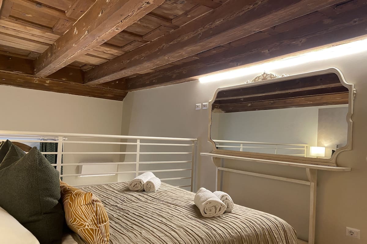 Un dormitori amb sostre de fusta, un capçal de llit blanc, un llit amb tovalloles arremolinades, un mirall i una tauleta de nit.