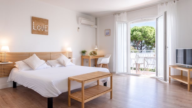 A modern bedroom with a wooden bed, a bedside table with a lamp, a small desk with a chair, and a TV on a stand. - Hotel & Studios ASTOR (Antibes)